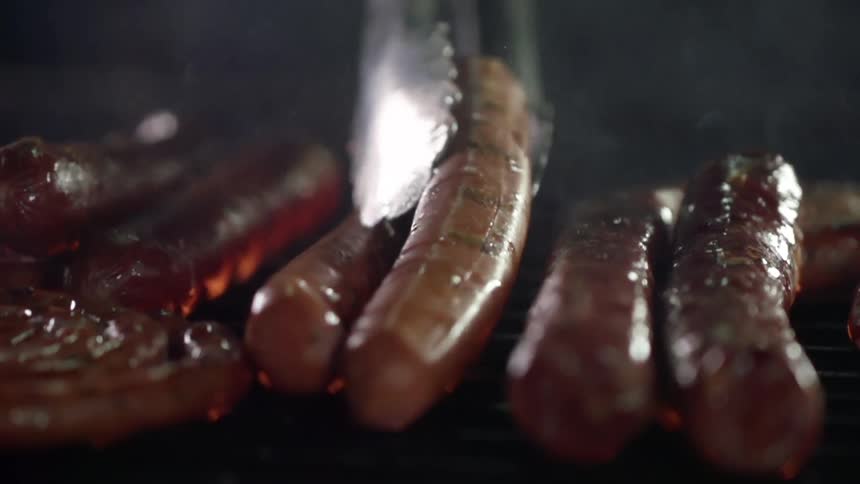 A person uses metal tongs to turn a hot dog on a grill