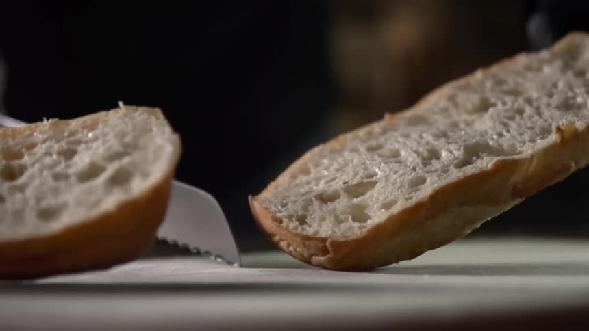 Gloved hands use a serrated knife to slice through a bread roll