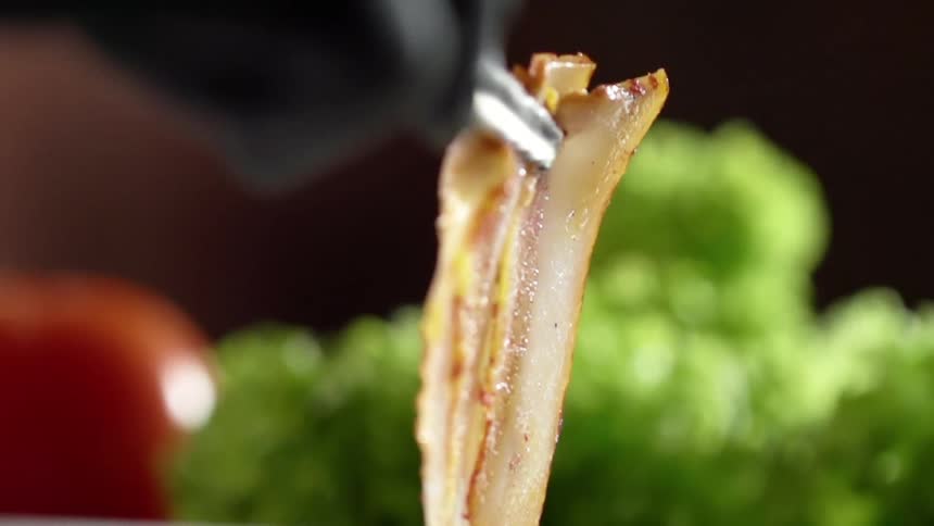 Chef wearing black latex gloves places a strip of cooked bacon on a white ceramic place using pliers