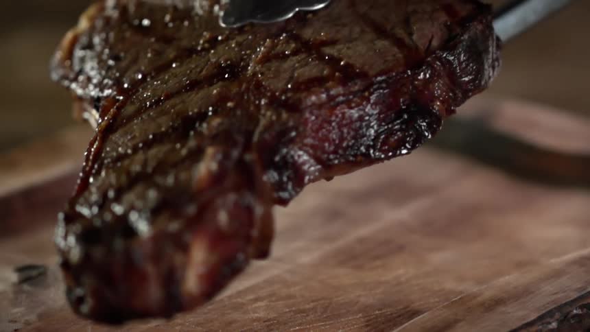 Close up of a grilled steak being put on a steak board by a pair of tongs
