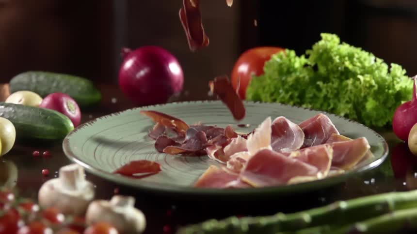 Slices of meat are dropped onto a plate that is surrounded with a variety of produce and mushrooms