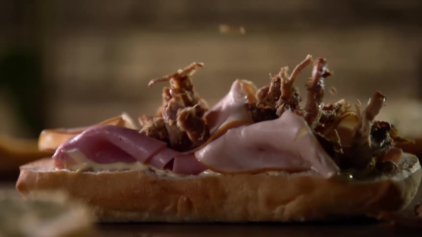 A sliced bread roll with sliced ham on it also has pulled pork sprinkled on top of it