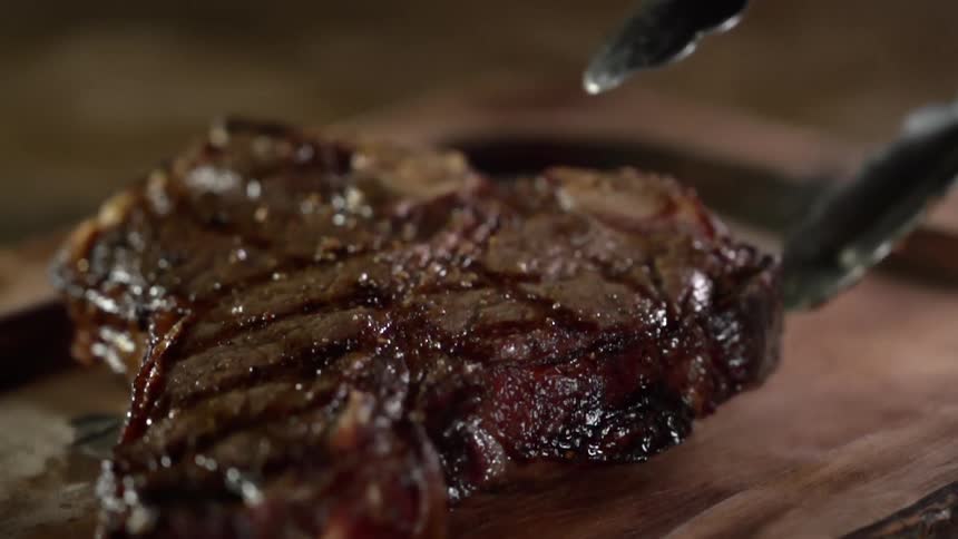 Grilled t bone steak being placed in a wooden board with pliers