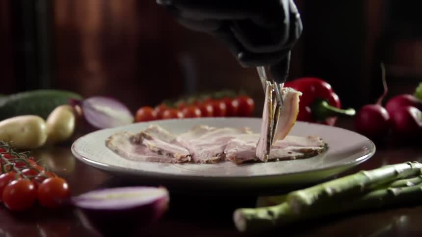 A gloved hand uses tongs to place sliced meat on a plate