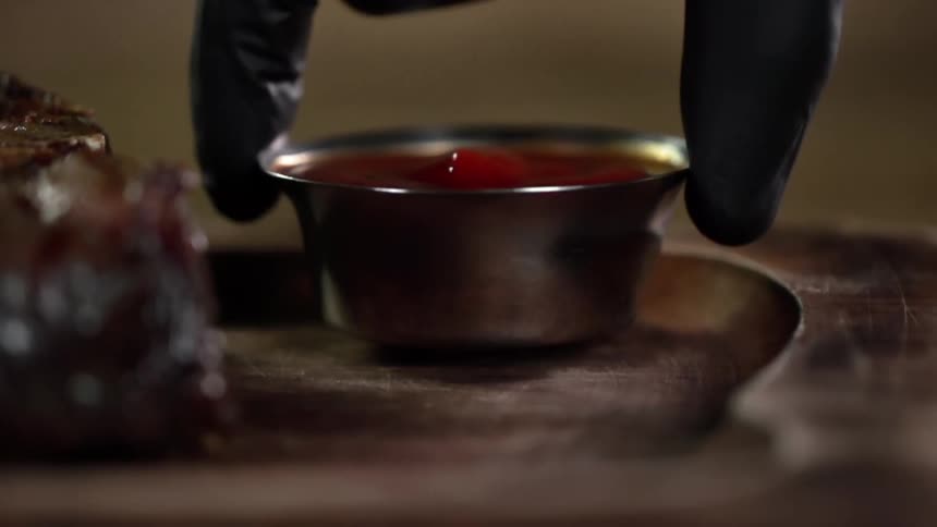 Chef wearing black latex gloves places a small metallic dish with ketchup next to steak in a wooden board
