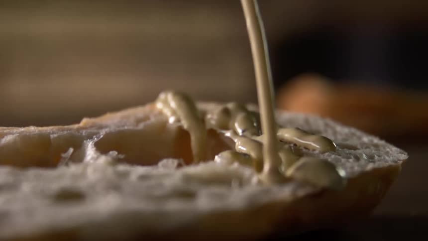A creamy yellow condiment is poured onto half of a bread roll