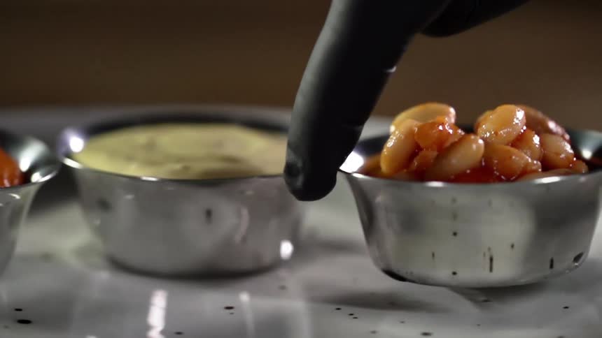A gloved hand places a small bowl of beans next to dipping sauces on a white plate