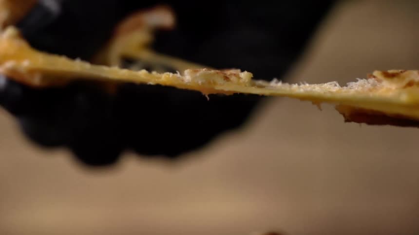 A person wearing black gloves pulls a piece of cheesy food apart