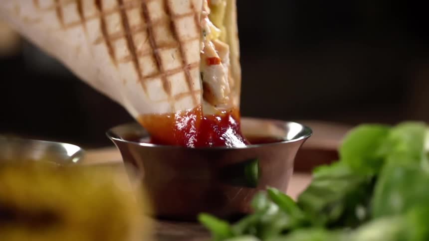 A grilled fajita is dipped into a small cup of sauce and lifted upwards as the sauce drips off of it