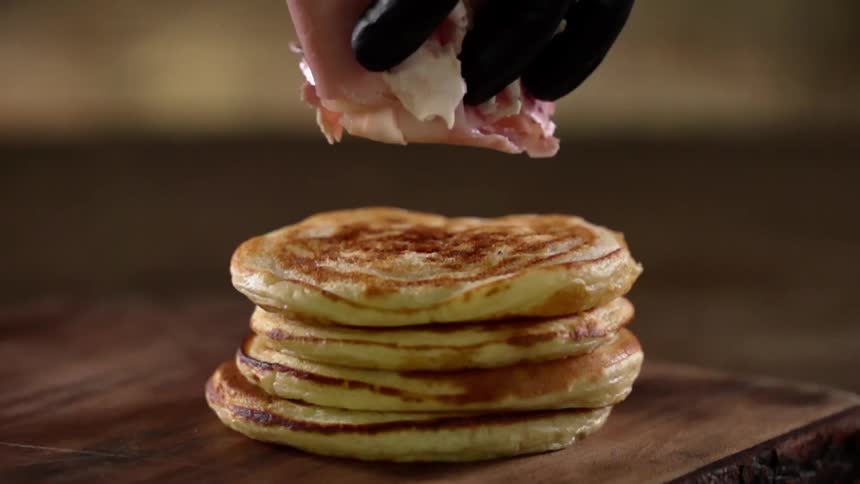 A black gloved hand drops ham on top of a stack of pancakes