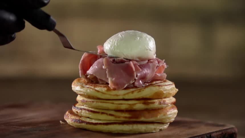 A poached egg is placed atop a pile of sliced ham which itself lies atop a stack of pancakes