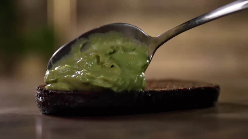 A spoon covered in guacamole taps against a piece of dark bread