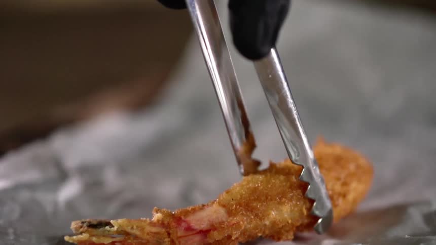 A person lowers a piece of breaded shrimp with metal tongs