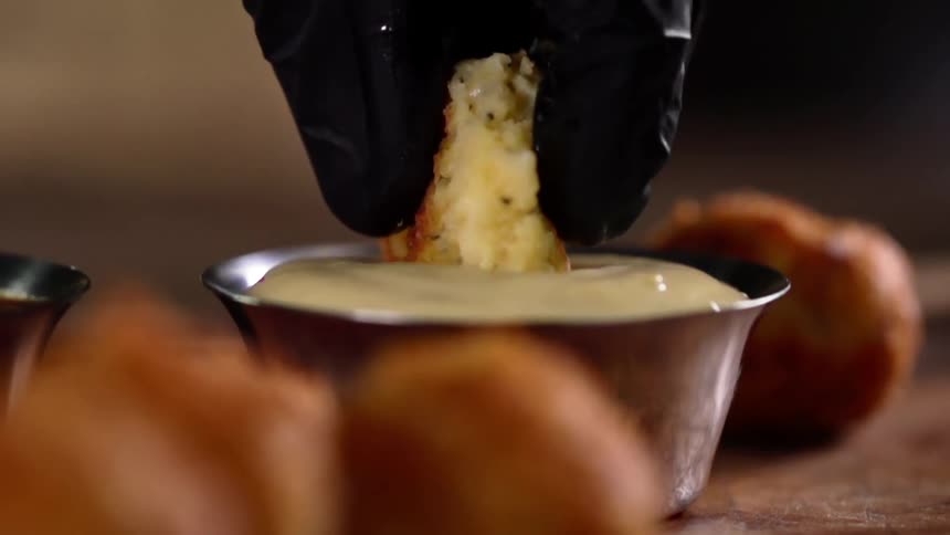 A person wearing a black glove dunks a piece of bread into dipping sauce