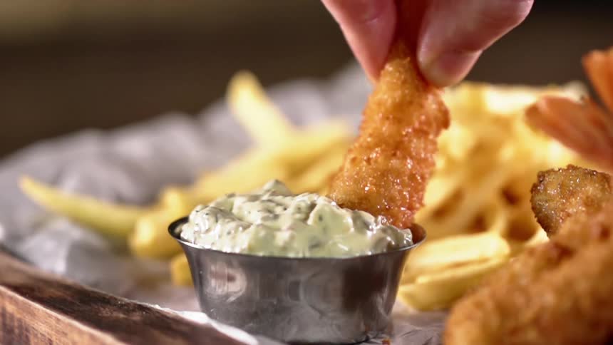 A white man dips a breaded shrimp into a small metal bowl filled with tartar sauce