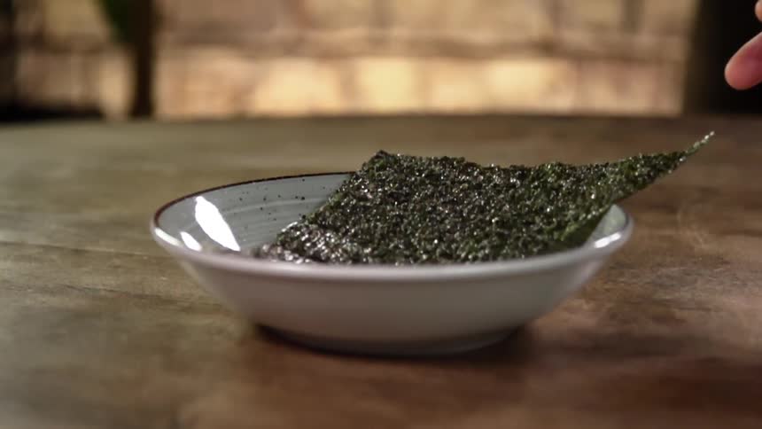 A hand drops a sheet of seaweed into a bowl on a table