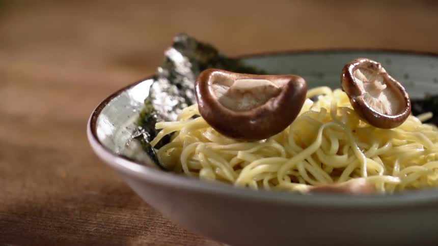 Mushroom caps drop onto pasta in bowl on wooden table