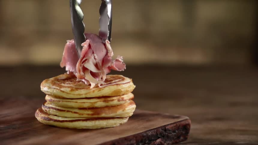 A pile of sliced ham is placed on top of a small stack of pancakes using a pair of metal tongs