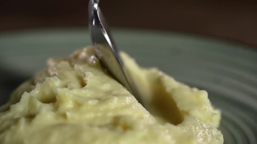A spoon moves through a pile of mashed potatoes on a plate