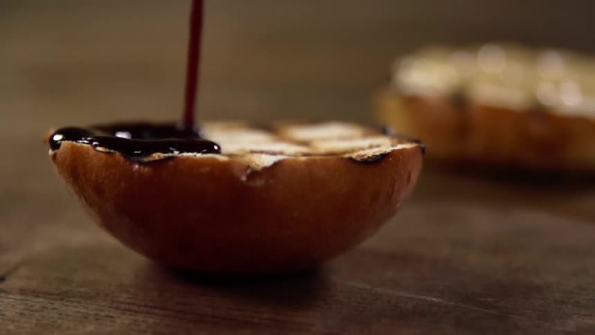 The open side of a grilled and sliced bread bun has a dark sauce slowly drizzled over its surface