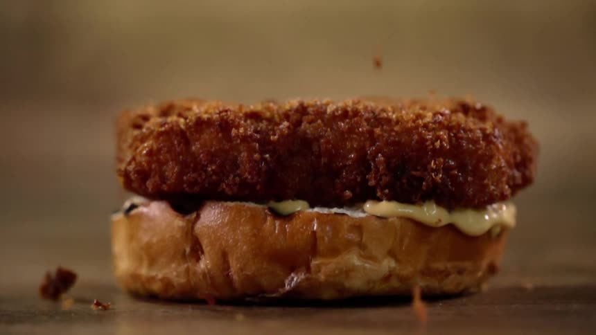 A breaded patty is dropped onto the bottom of a bun with a creamy sauce