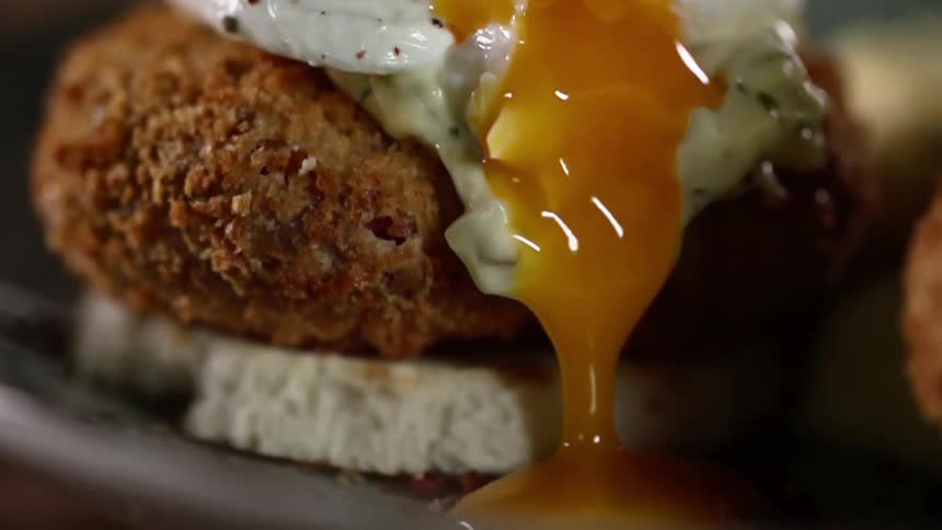 A runny egg sitting on top of a crab cake has its yolk slowly ooze down onto the plate they are on