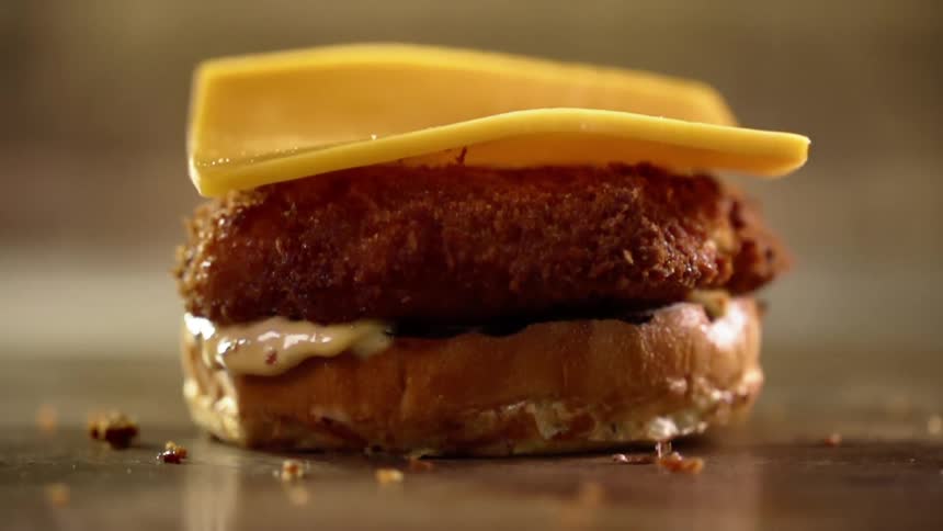 A single slice of yellow cheese is dropped on top of a breaded meat patty sitting atop a sliced bun