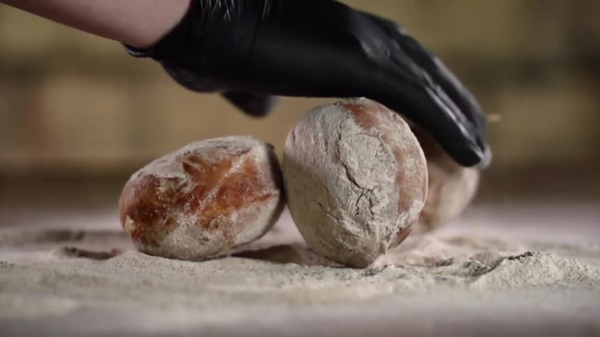A person wearing a black disposable glove pushes three baked rolls around in a pile of flour