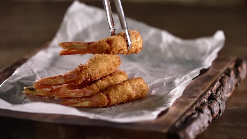 A pair of metal tongs drops a piece of breaded shrimp onto a piece of paper