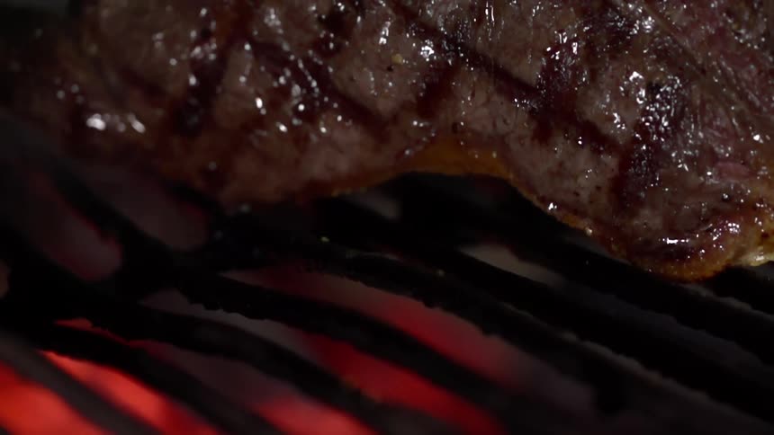 A moist slab of grilled steak is being lifted from the flame grill using a pair of tongs