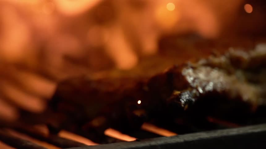 A slab of steak sits atop a grill as an open flame burns in the background