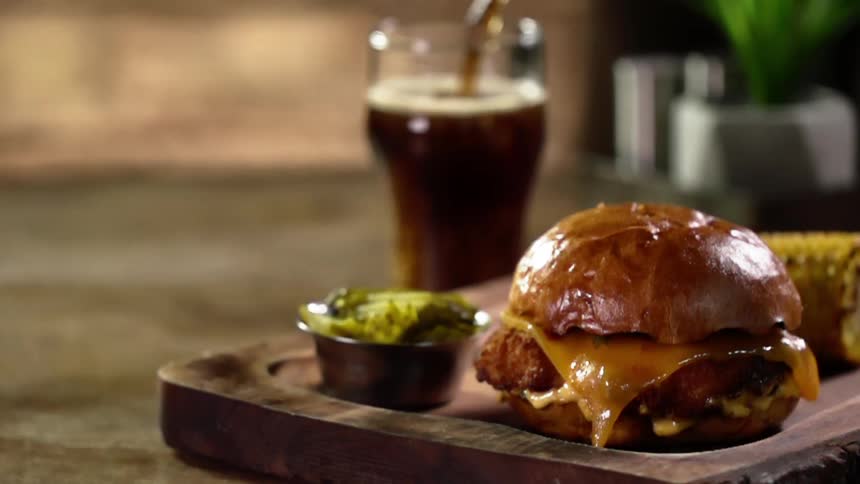 A glass fills with a drink behind a platter with a burger and corn cob