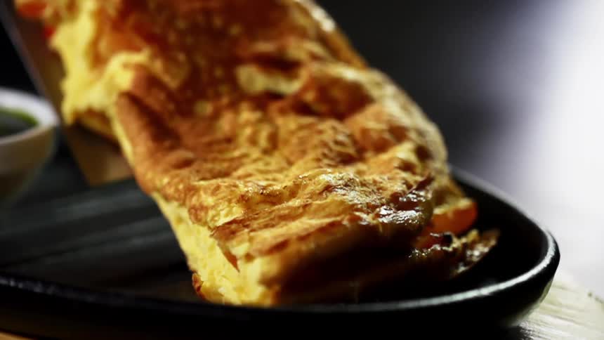 A browned omelet slides off a spatula onto a black tray next to a dipping bowl filled with dark colored sauce
