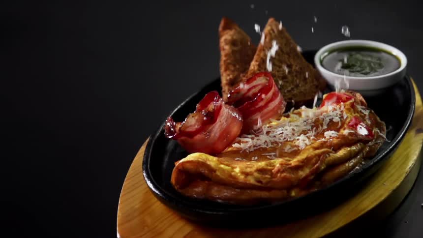 Cheese is sprinkled onto an omelet next to bacon and toast
