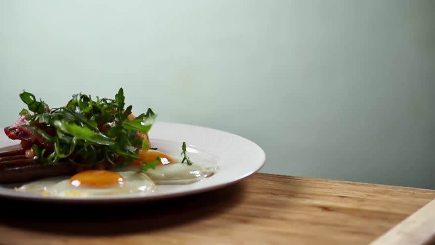 Arugula leaves falling over a white ceramic plate with eggs and bacon breakfast