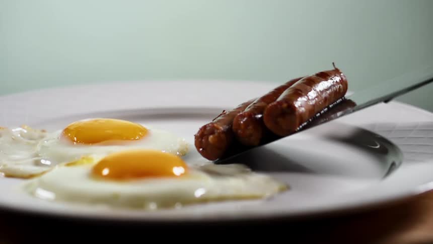 A trio of small sausages is placed onto a white plate next to two fried eggs