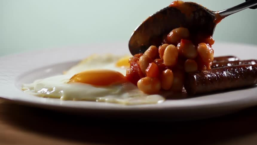 Baked beans are spooned onto a plate with eggs and sausage