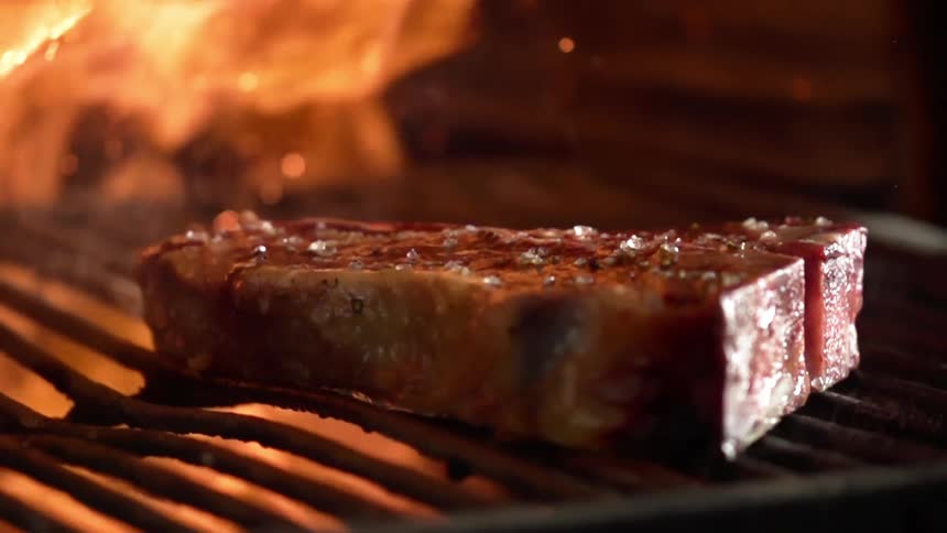 A pair of metal tongs places a steak on a grill