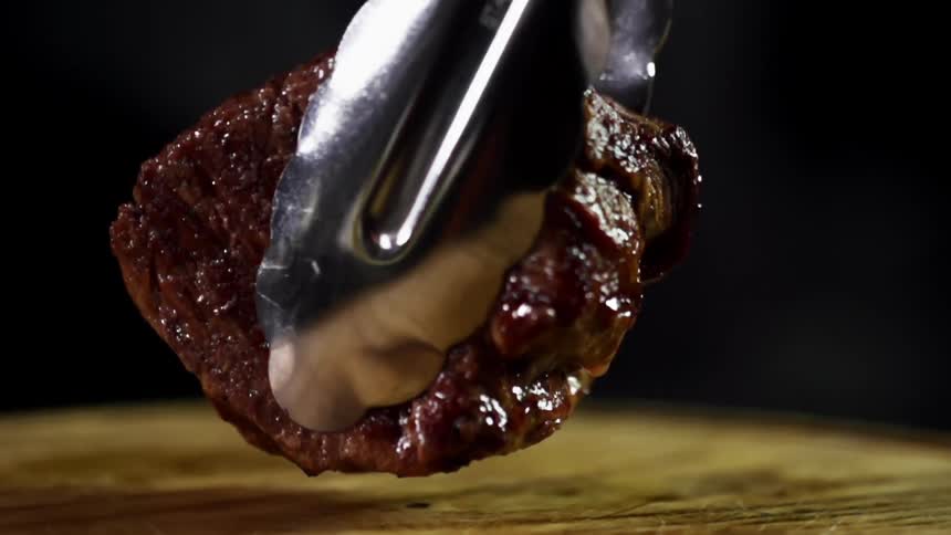 A small steak being placed on a circular wooden board with a pair of tongs