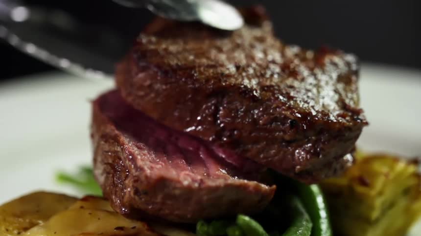 Tongs place a piece of steak on top of another steak on a dinner plate