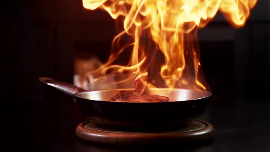 A pan that contains a steak is ignited with a hand torch in order to sear the steak