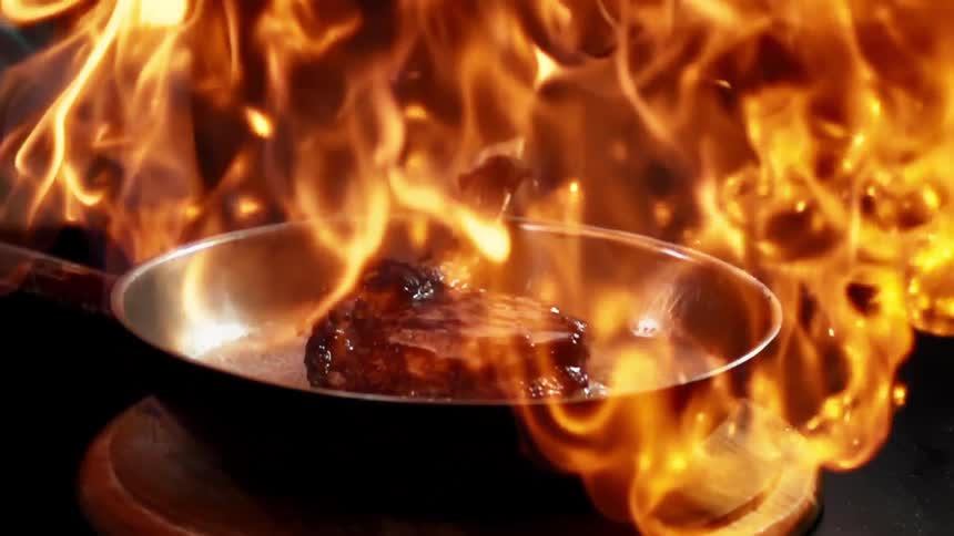 A thick piece of steak is being cooked in a large metal pan and is engulfed in a large flame