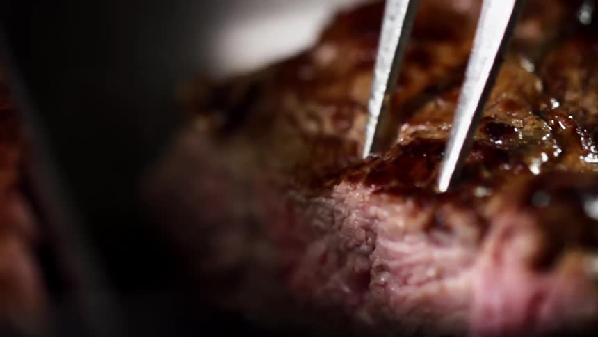 A moist piece of grilled steak is cut with a knife and slid apart using a fork