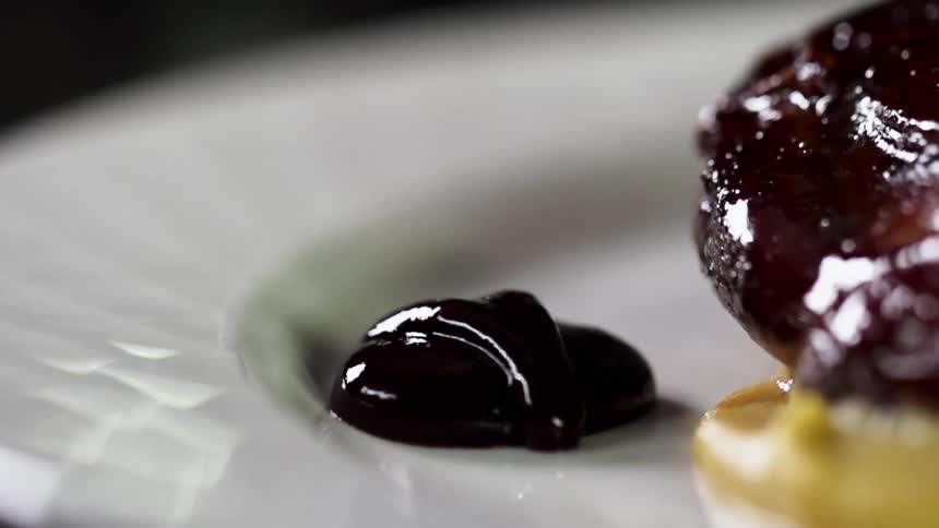 Dark thick sauce being poured over a white ceramic dish next to glazed food