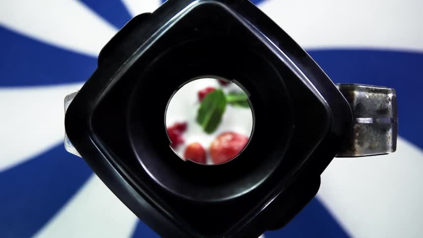 A person places the lid onto the top of a blender containing strawberries and green spinach leafs suspended in white cream