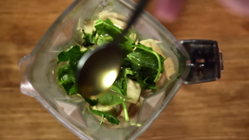 A spoonful of honey is held above a blender and the honey is drizzled over the leafy spinach inside