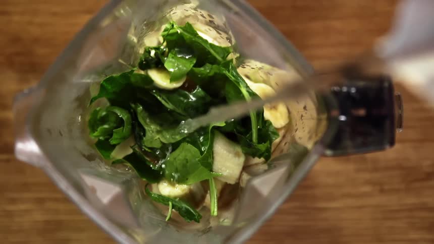 Pouring water from a jar into a mixer filled with leafy greens and bananas
