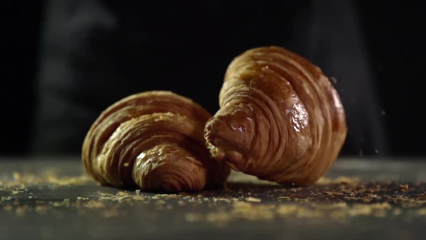 A baked croissant is dropped onto another one and small flakes of the croissants surface fall to the counter top surface