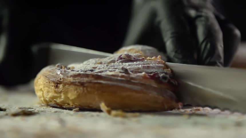 A person wearing black disposable gloves slowly cut through a pastry using a long bread knife