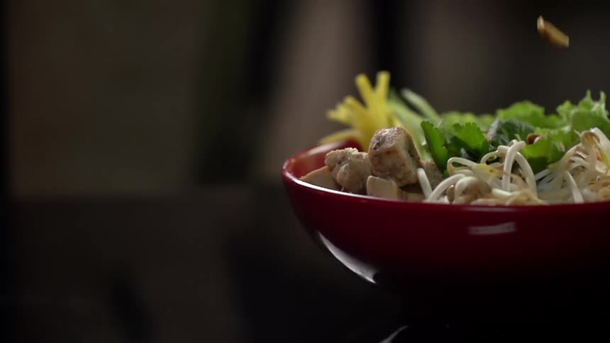 Nuts falling into a red bowl containing chopped chicken vegetables and noodles